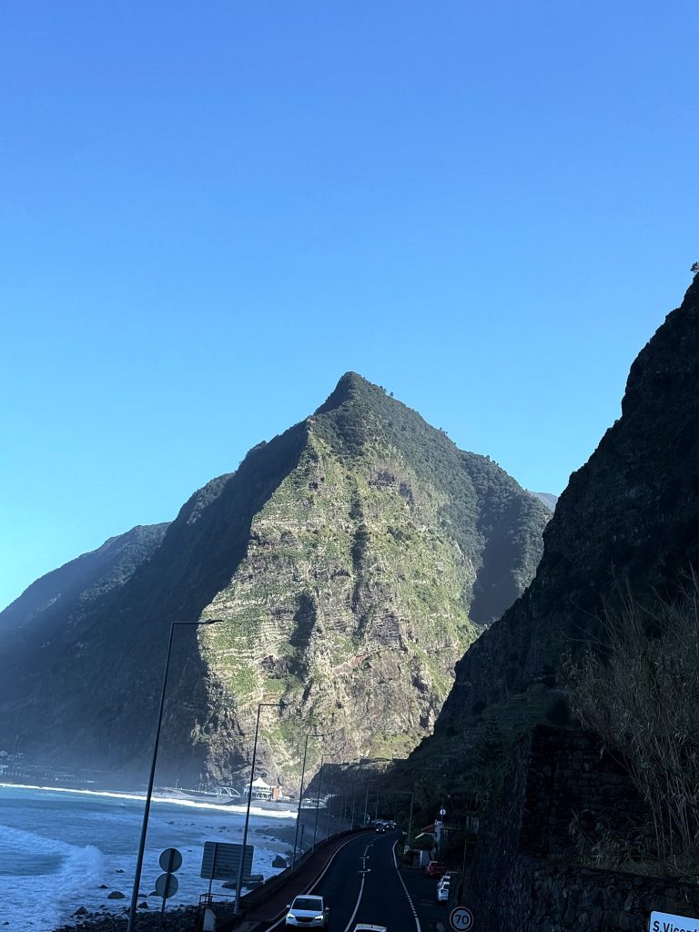 A pyramid-shaped peak above the coast road and the ocean, with a 70 speed limit sign and a São Vicente name sign by the road