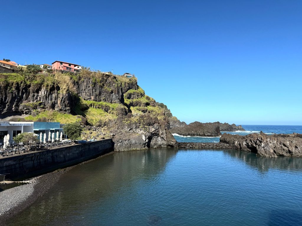 A calm natural pool below a rocky cliff with houses on top, a pavilion with parasols by the pool, the ocean in the background