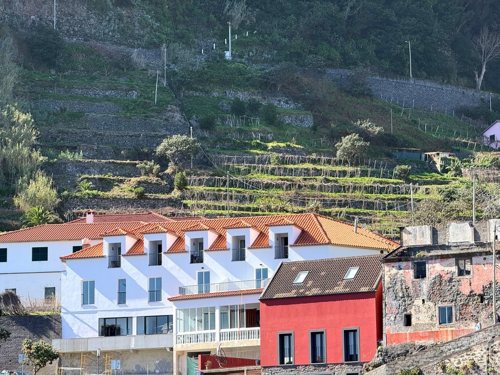 A white building with an orange roof, a red house and an old building with peeling walls below terraces on the slope