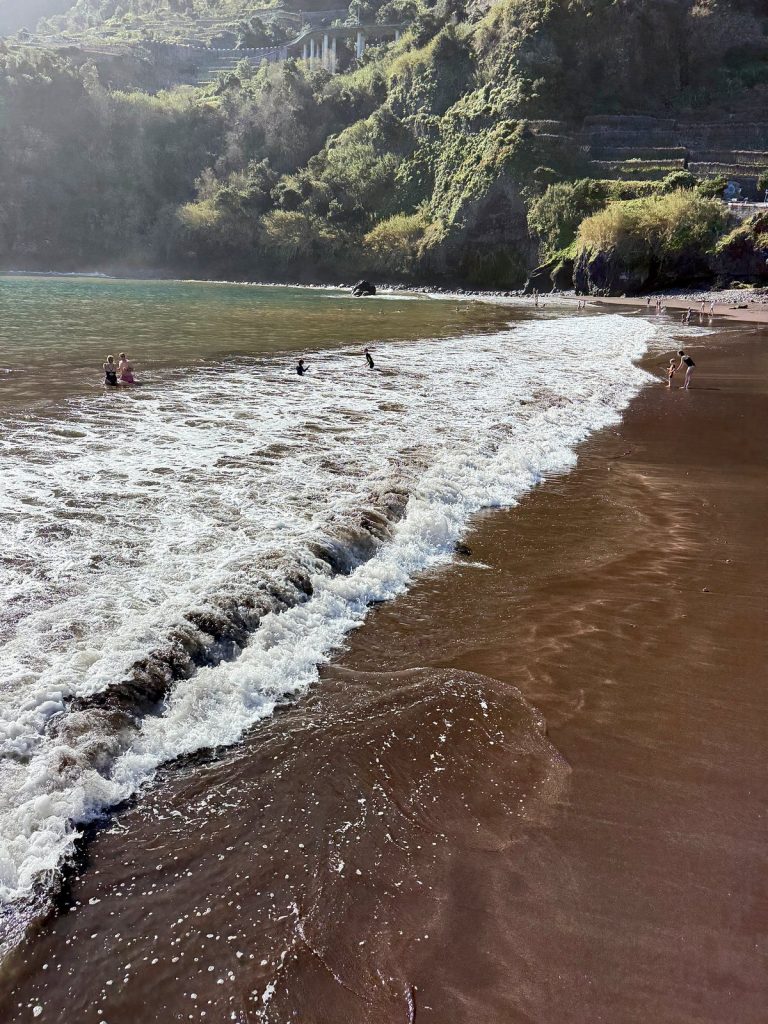 A foaming wave running up the dark sand, with green cliffs in a light haze and a few people in the water behind