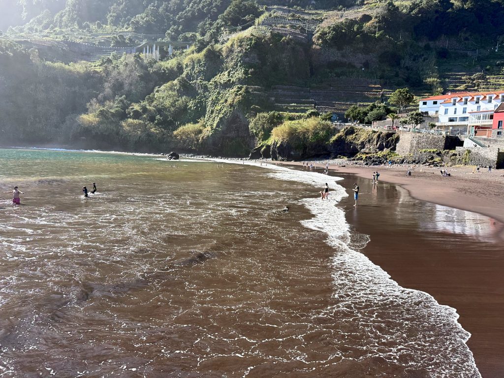 Waves rolling onto a dark-sand beach, with green slopes and white houses behind and small figures of swimmers in the water