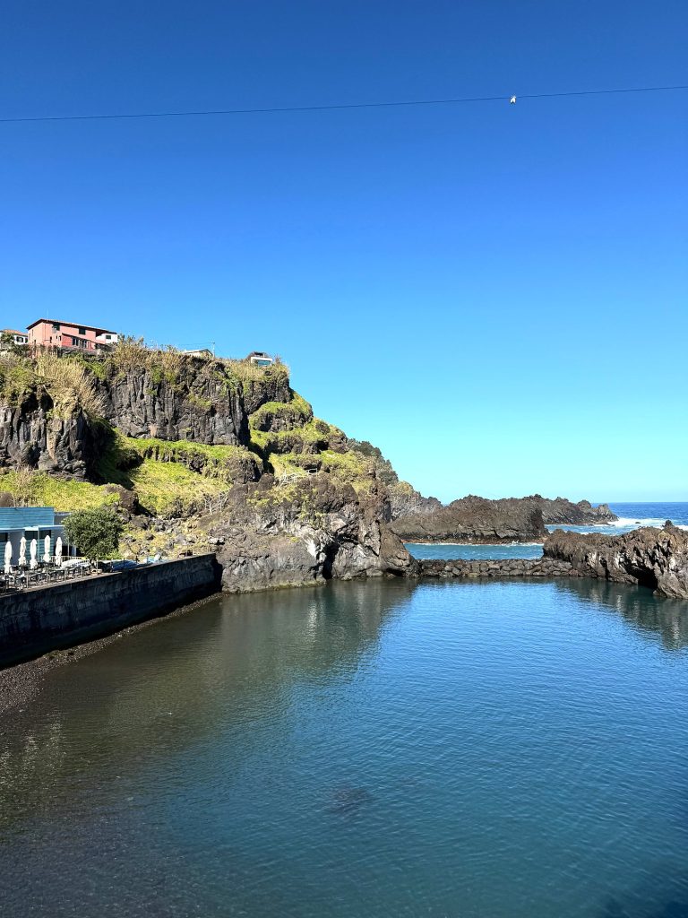 A natural pool with calm water between volcanic rocks, a cliff with a red house above it and restaurant parasols on the left