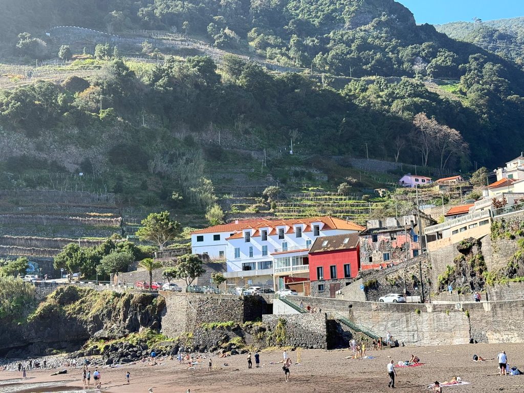 Dark-sand beach below a green terraced slope, a white building with an orange roof and a red house by the shore, small figures on the beach