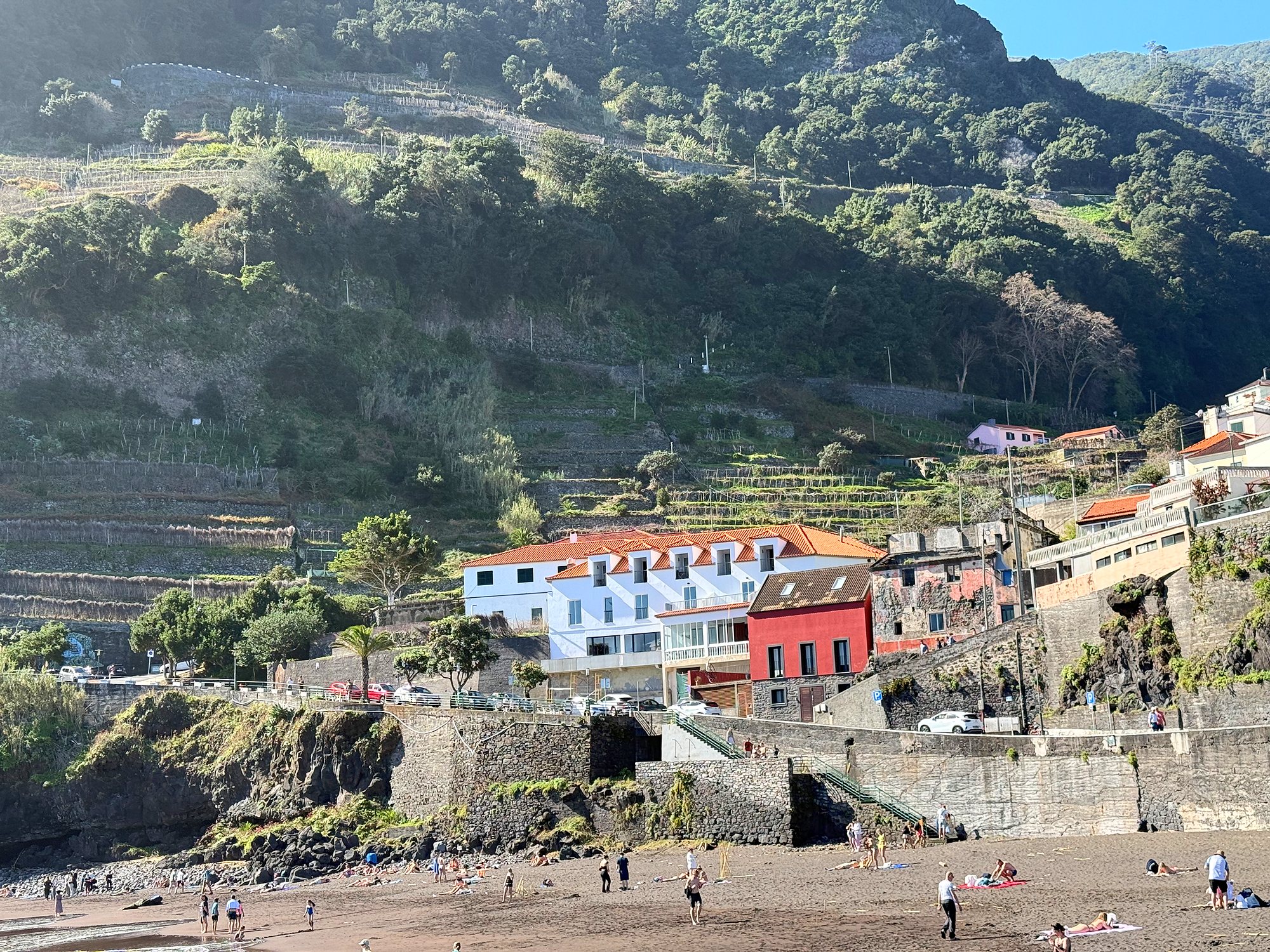 Dark-sand beach below a green terraced slope, a white building with an orange roof and a red house by the shore, small figures on the beach