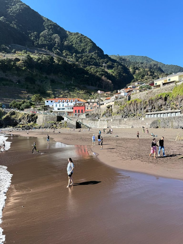 People walking on a dark-sand beach, with a white house and a red house, harbour walls and green terraced slopes behind