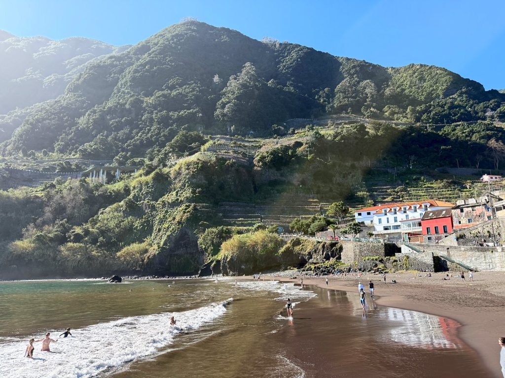 A dark-sand beach below a steep green terraced mountain, people swimming in the foaming waves by the shore, a white house and a red house on the right