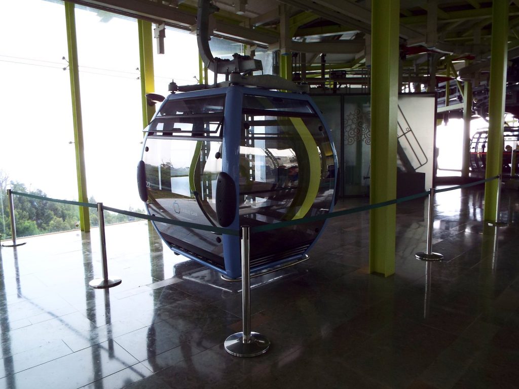An empty blue-grey cable car cabin in the station, with the green hillside behind the glass